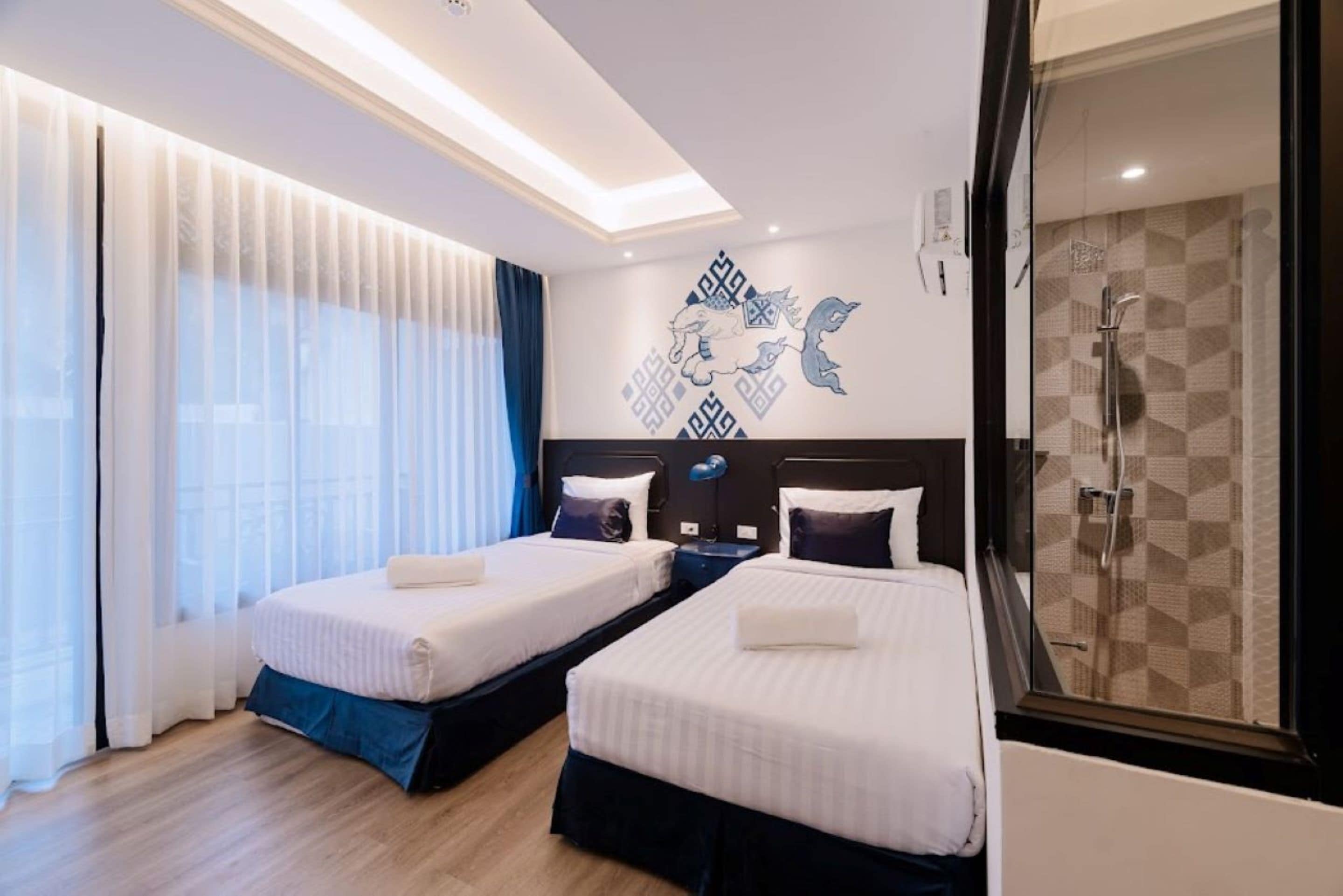 Habitació d'hotel amb dos llits, una finestra gran amb cortines translúcides, una obra d'art moderna a la paret i un bany amb un disseny de rajoles geomètric.