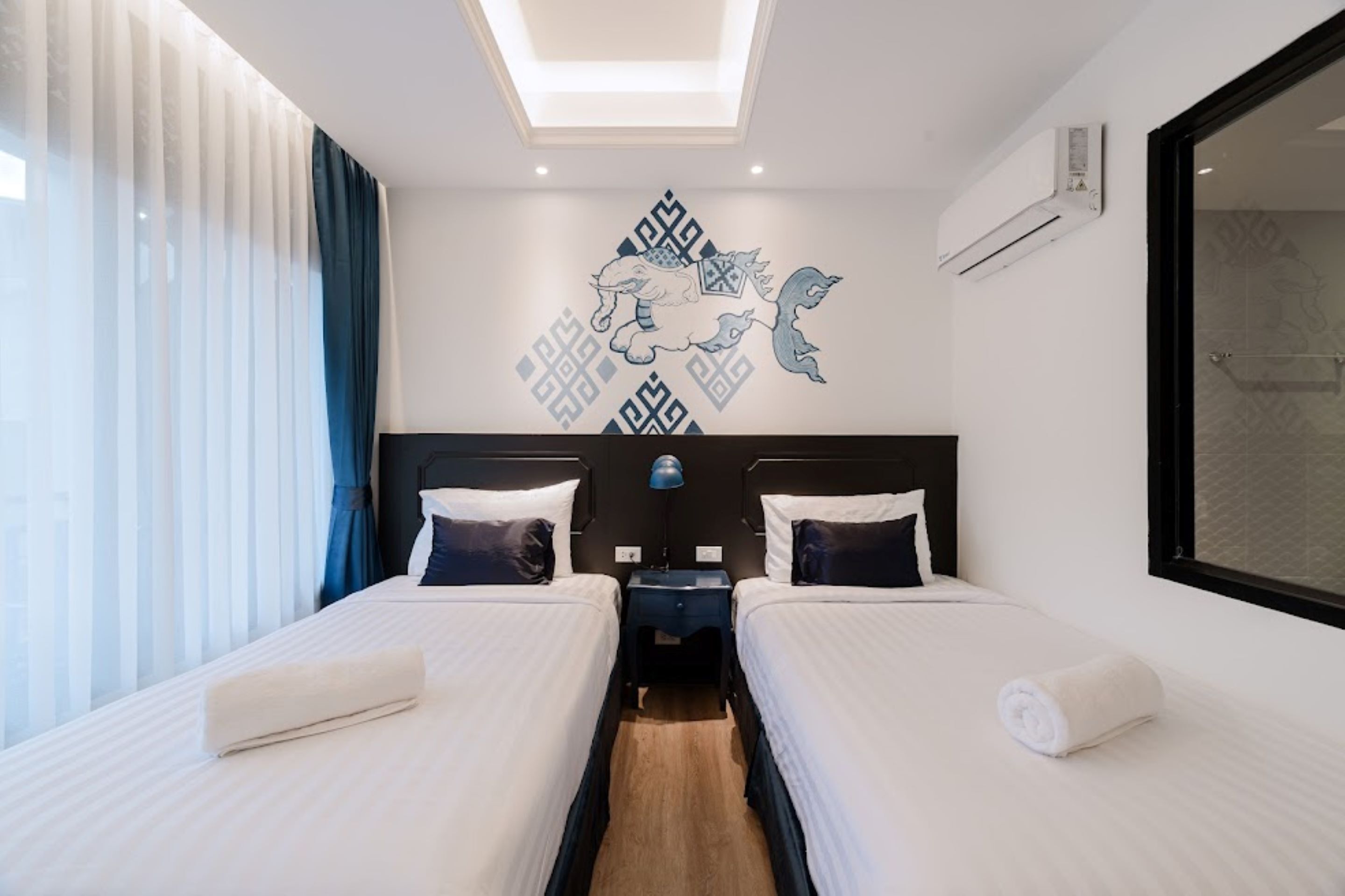 Habitación de hotel con dos camas, una lámpara en el techo, aire acondicionado y un amplio ventanal con cortinas.