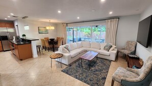 A spacious living area with a sofa, coffee table, and chairs, a kitchen with a refrigerator and dining set, and large windows offering a view of the outdoors. - Paradise 4 min to Beach with Heated Pool and Spa (Deerfield Beach)