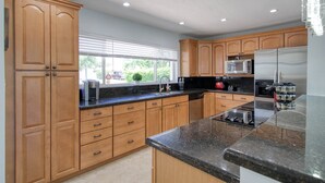 A modern kitchen with wooden cabinets, a granite countertop, stainless steel appliances, and a window with blinds. - Paradise 4 min to Beach with Heated Pool and Spa (Deerfield Beach)