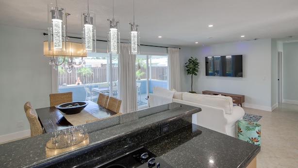A modern kitchen with a granite countertop, a dining area with wicker chairs, a white sofa, and a TV mounted on the wall. - Paradise 4 min to Beach with Heated Pool and Spa (Deerfield Beach)