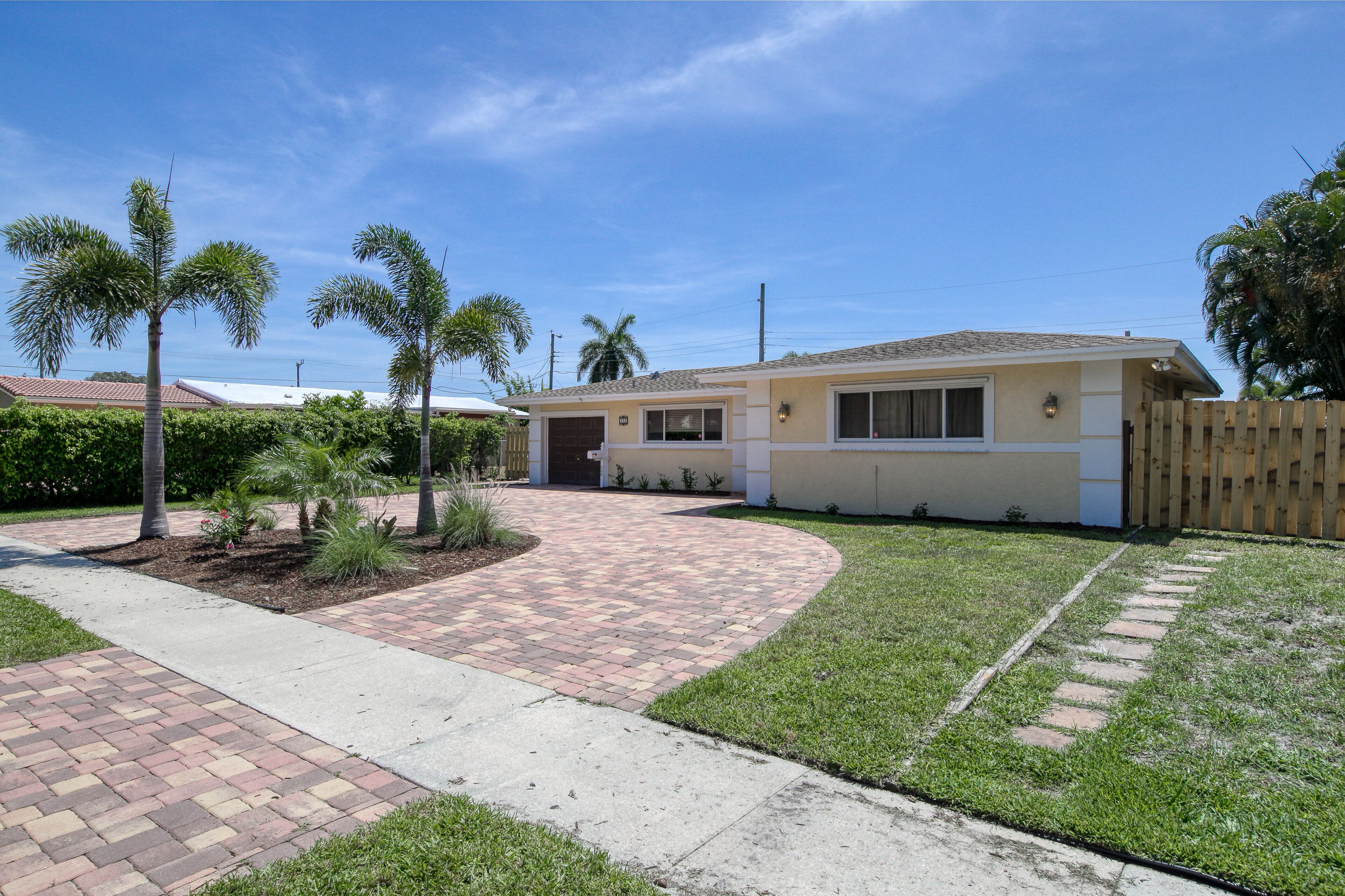 A single-story house with a paved driveway, a wooden fence, and palm trees.