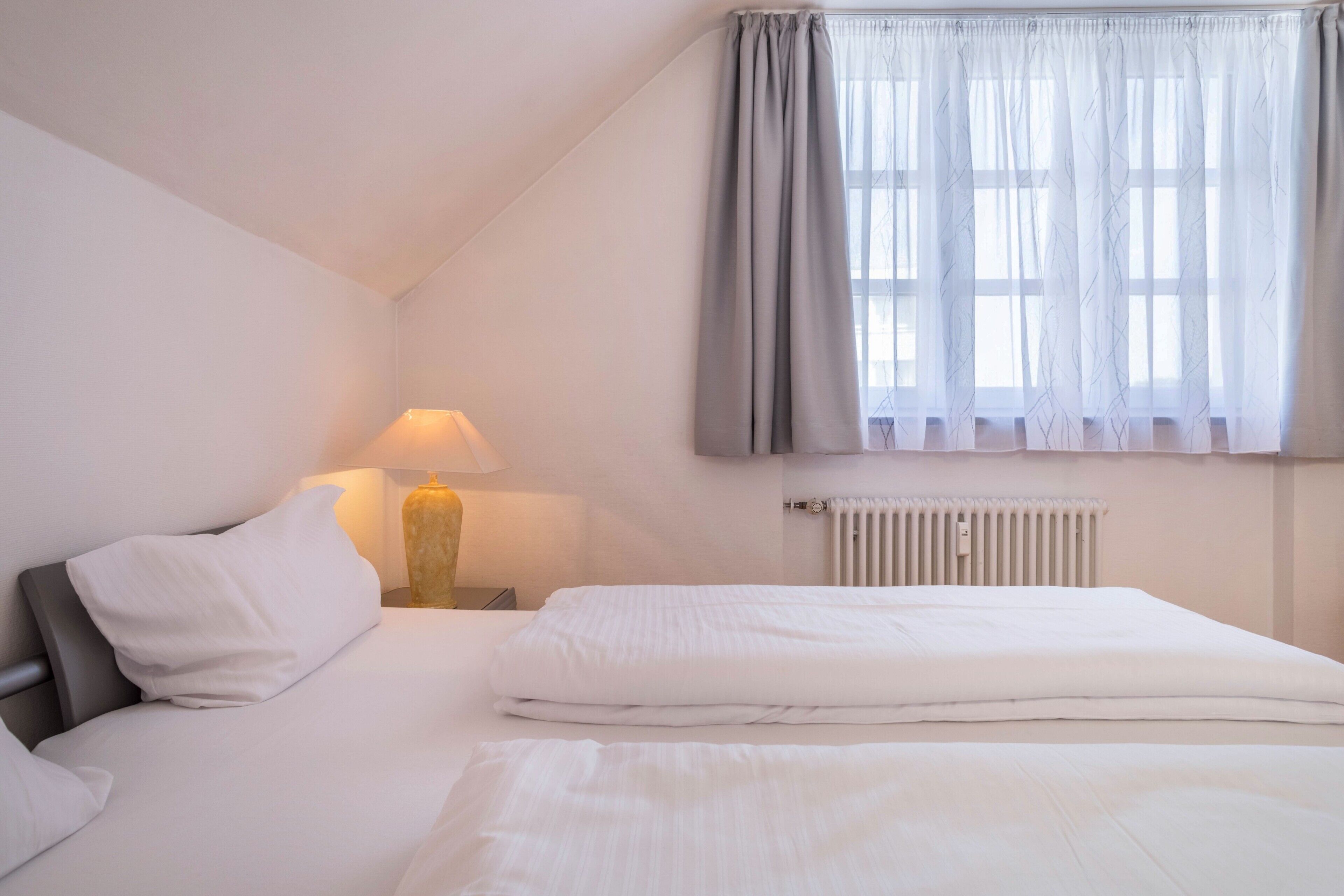 Une chambre à coucher avec un lit, une table de chevet, une lampe, un radiateur et une fenêtre avec des rideaux.