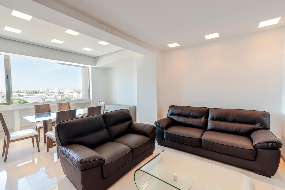Una sala de estar moderna con dos sofás de cuero marrón oscuro, una mesa de centro de vidrio y un comedor con mesa y sillas de vidrio.