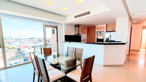 A modern kitchen with a glass dining table and chairs, a city view through large windows, and a spacious layout.