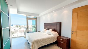 A modern bedroom with a large bed, a wooden nightstand, and a view of the city through a large window.