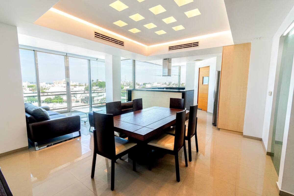 Un área de comedor moderna con una mesa grande y sillas, ventanas amplias que ofrecen vista a la ciudad, y una cocina con estufa y microondas.