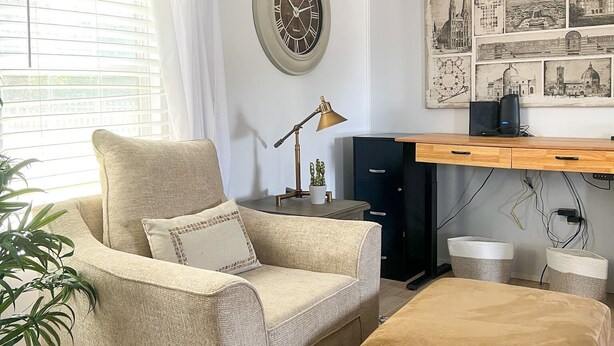 A cozy living room with a beige sofa, a small ottoman, a side table with a lamp, and a wall clock. - The Cozy Retreat - 3BR - Sleeps 7 - Near Greenville Mauldin & Simpsonville (Greenville)