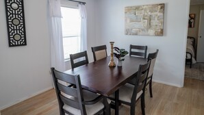 A dining area with a dark wooden table, six chairs, a decorative wall piece, and a window with white curtains. - The Cozy Retreat - 3BR - Sleeps 7 - Near Greenville Mauldin & Simpsonville (Greenville)
