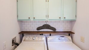 A laundry room with two front-loading washing machines, a wooden shelf, and a sign indicating it's open 24 hours. - The Cozy Retreat - 3BR - Sleeps 7 - Near Greenville Mauldin & Simpsonville (Greenville)