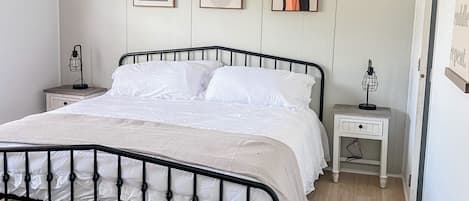 A neatly made bed with a black metal frame, white bedding, and a patterned rug. There are two bedside tables with lamps, and three framed artworks on the wall.