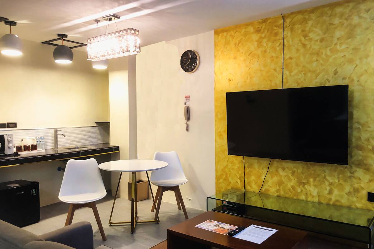 Un salon moderne comprenant un canapé, une table basse, un coin repas avec une table et des chaises, un téléviseur à écran plat fixé à un mur texturé, et un coin cuisine avec un évier et des appareils électroménagers posés sur le plan de travail.