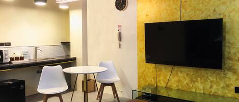 A modern living room with a sofa, a coffee table, a dining area with a table and chairs, a flat-screen TV mounted on a textured wall, and a kitchen area with a sink and countertop appliances.