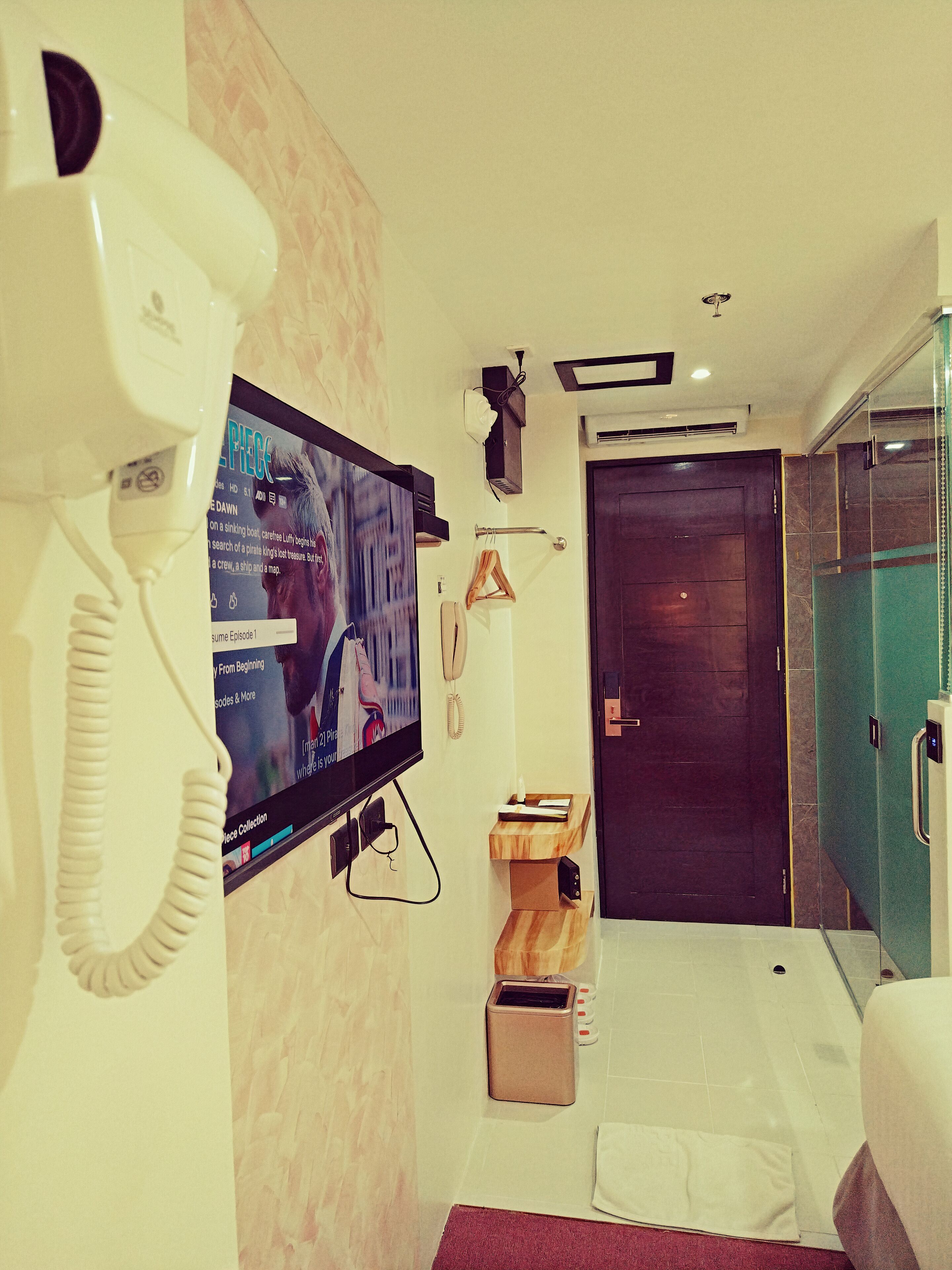 Una camera d'albergo con televisore, telefono, un piccolo ripiano in legno, una porta e un box doccia in vetro.