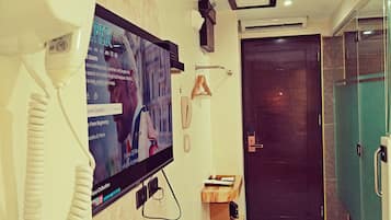 Una camera d'albergo con televisore, telefono, un piccolo ripiano in legno, una porta e un box doccia in vetro.