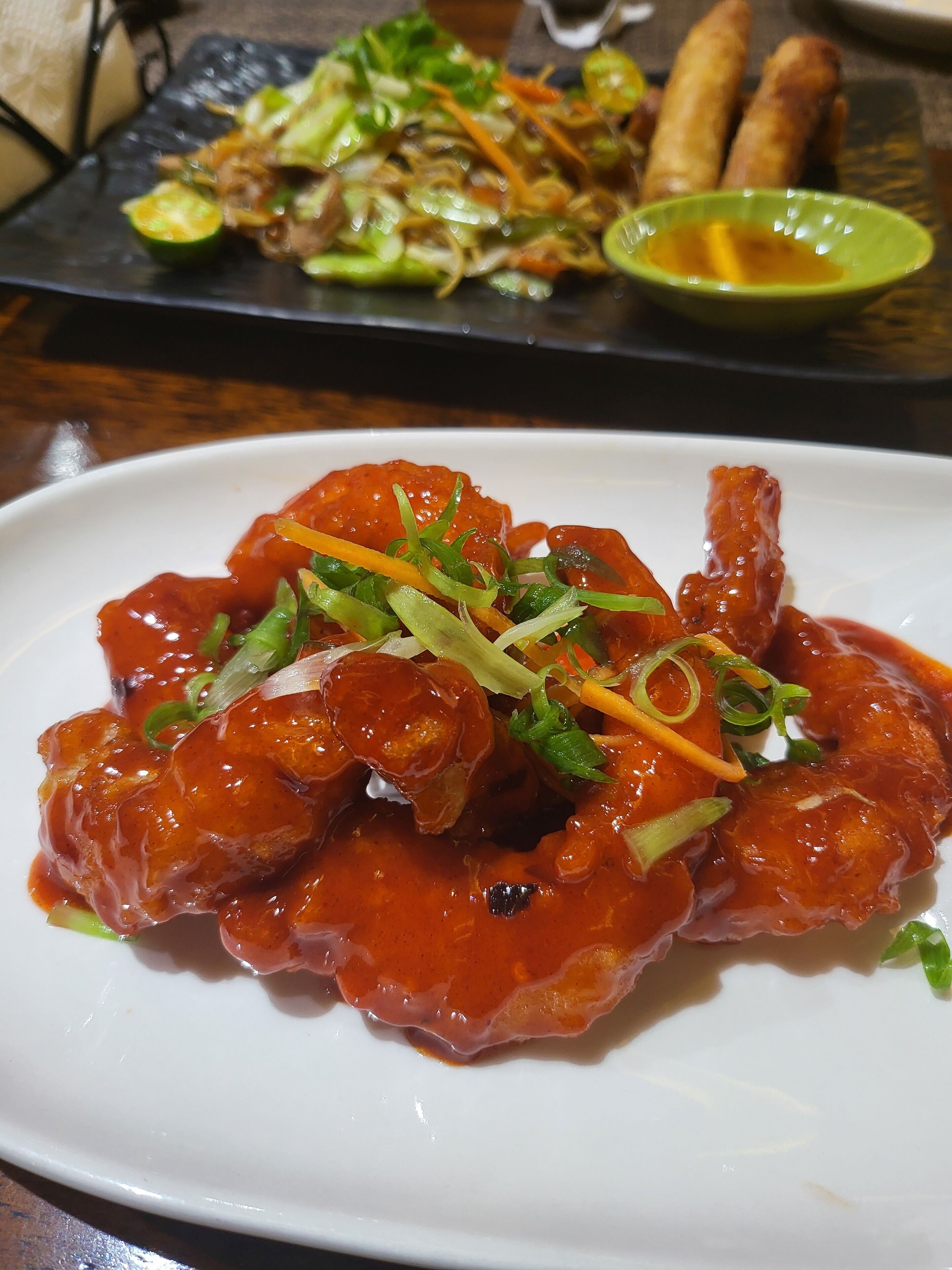 A plate of sweet and sour chicken with green onions and carrots, and a side of vegetables and spring rolls on a table.