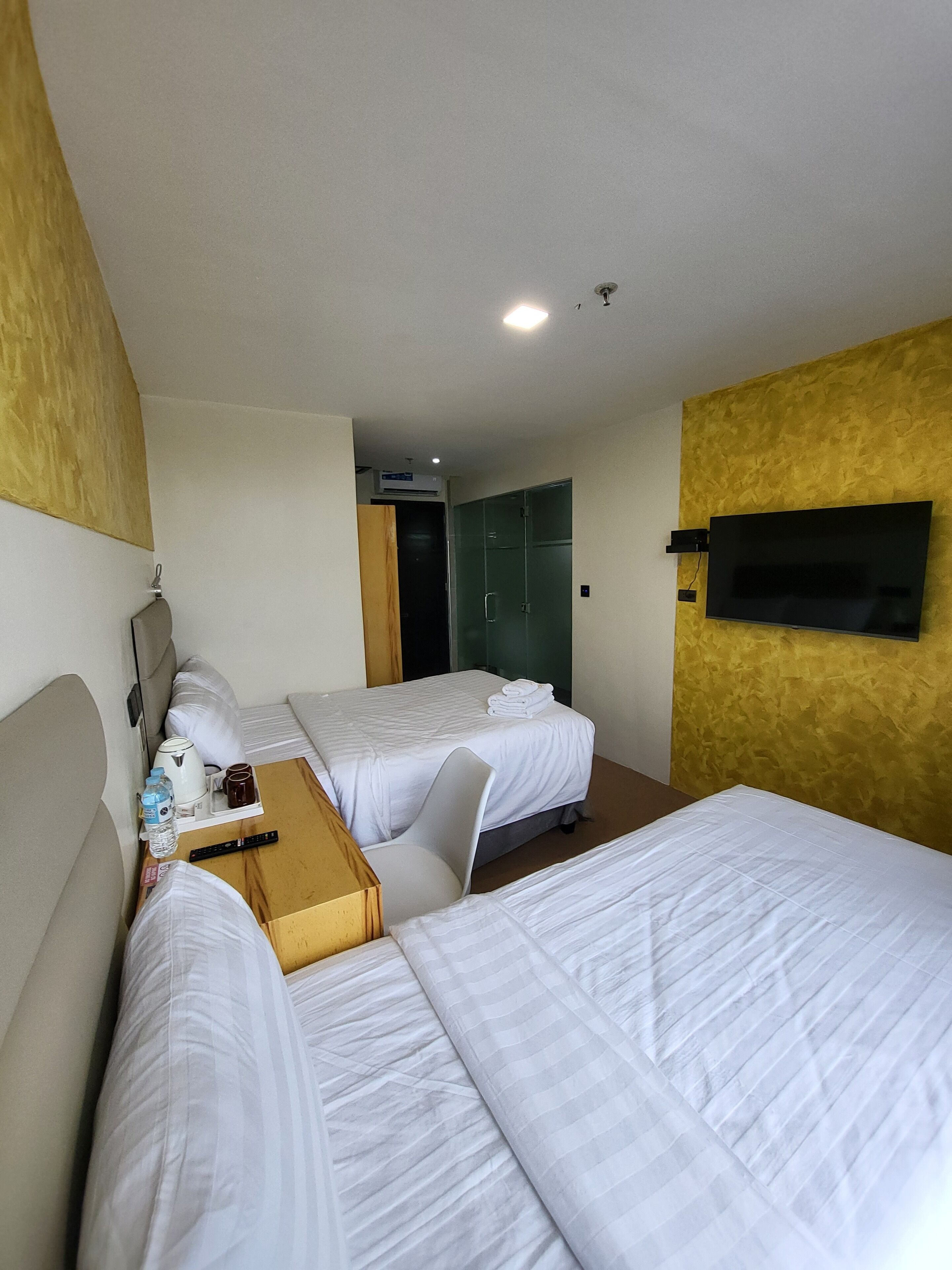 A hotel room with two beds, a TV mounted on the wall, a wooden side table with a kettle and cups, and a door leading to a bathroom.