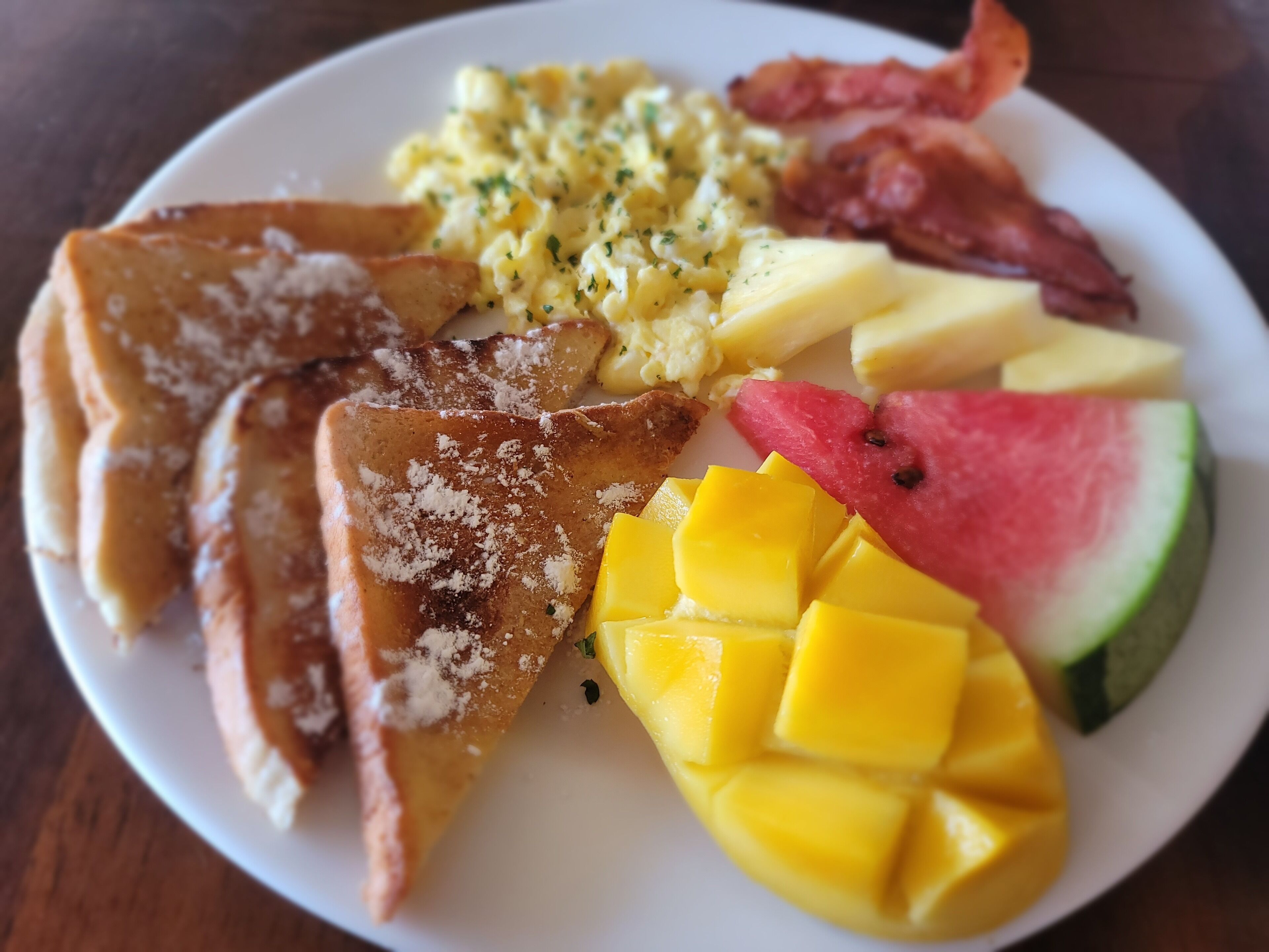 A plate with toast, scrambled eggs, bacon, and fruit including watermelon and mango.