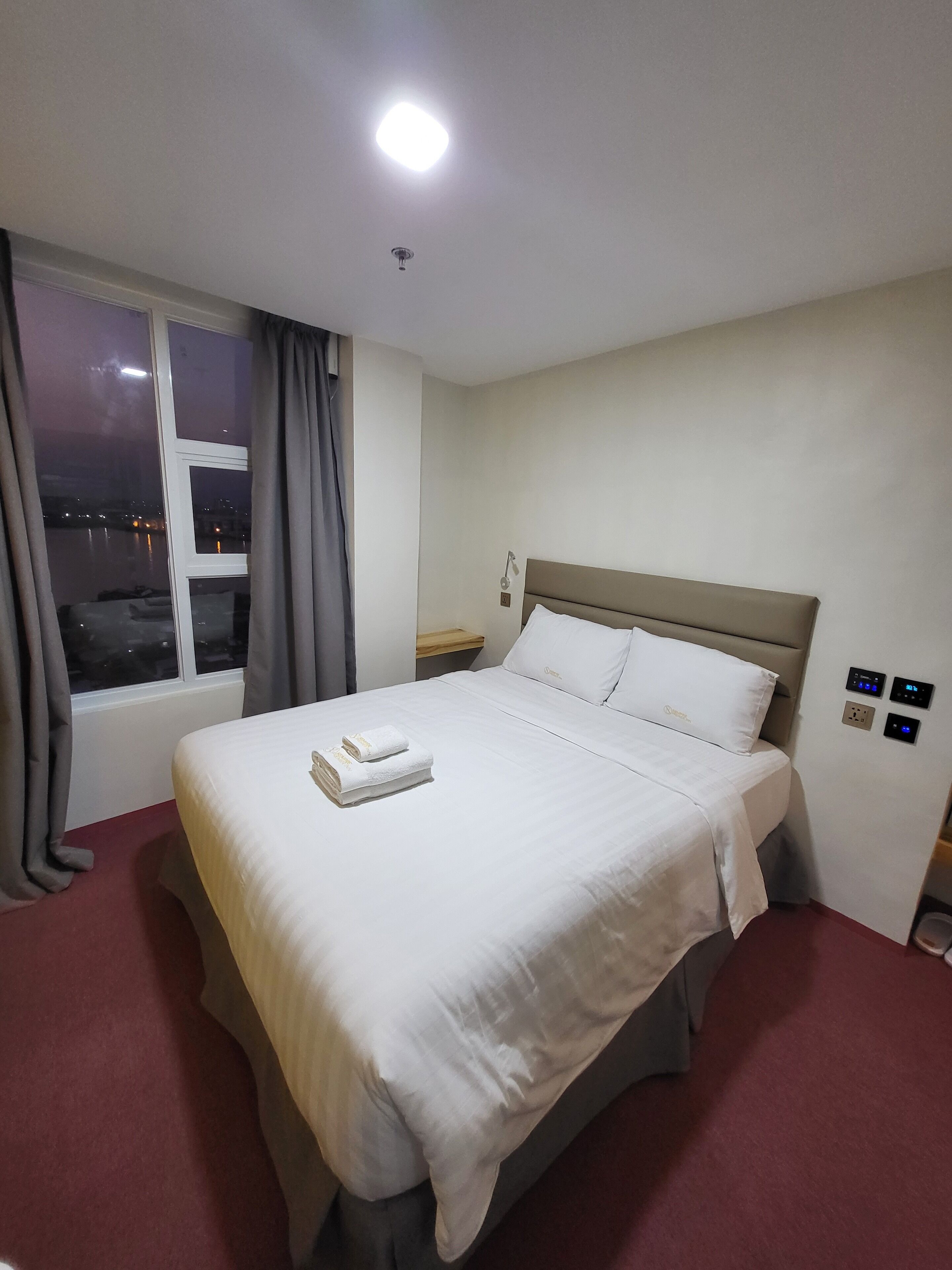 Hotellihuone, jossa on suuri sänky, yöpöytä, verhoilla varustettu ikkuna ja näkymä kaupunkiin yöllä.