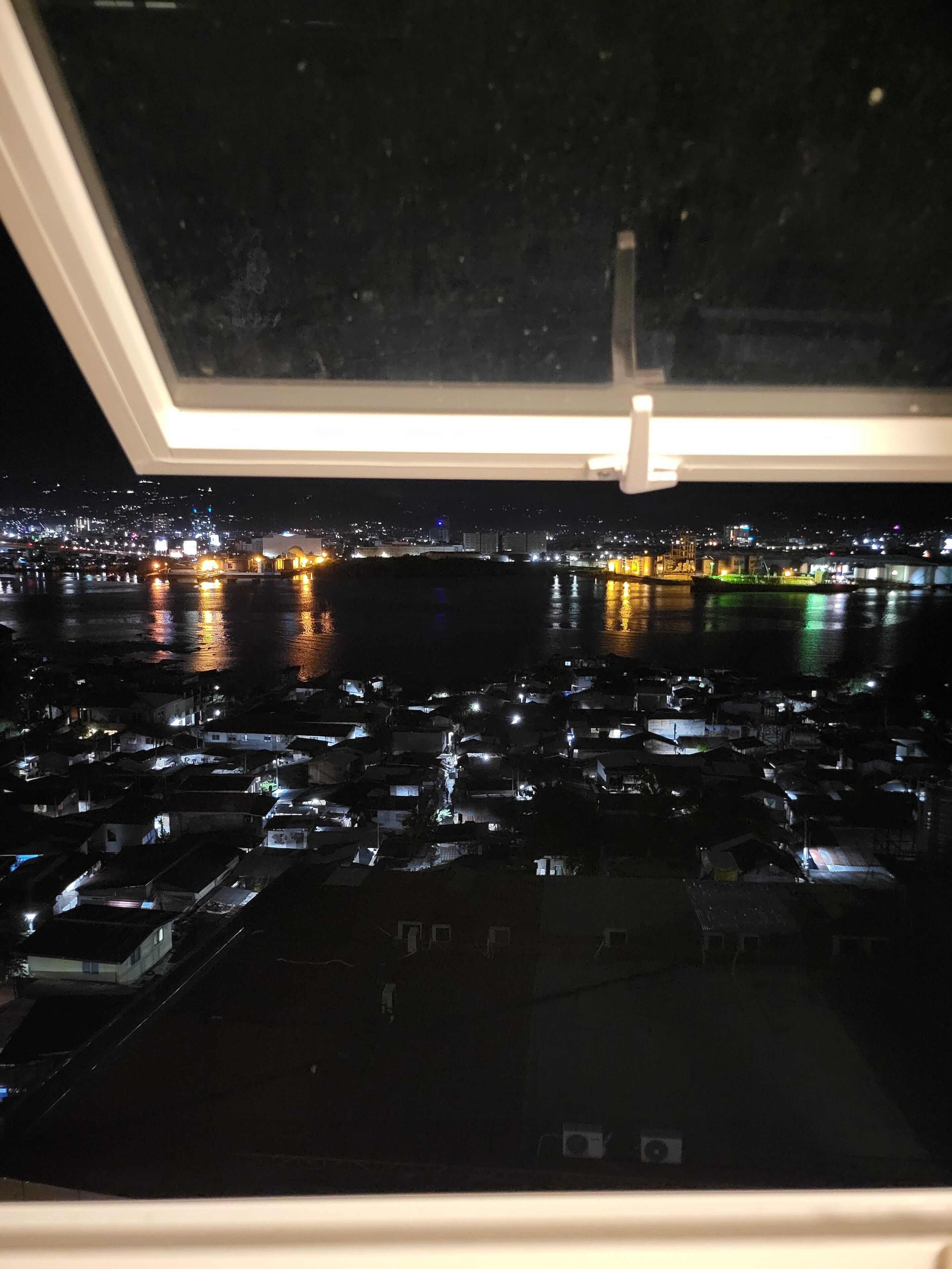 Vista nocturna desde una ventana con vista a un paisaje urbano con edificios iluminados y una masa de agua.