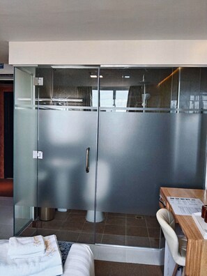 A modern hotel room with a glass-enclosed shower area, a wooden desk, and a bed with towels.