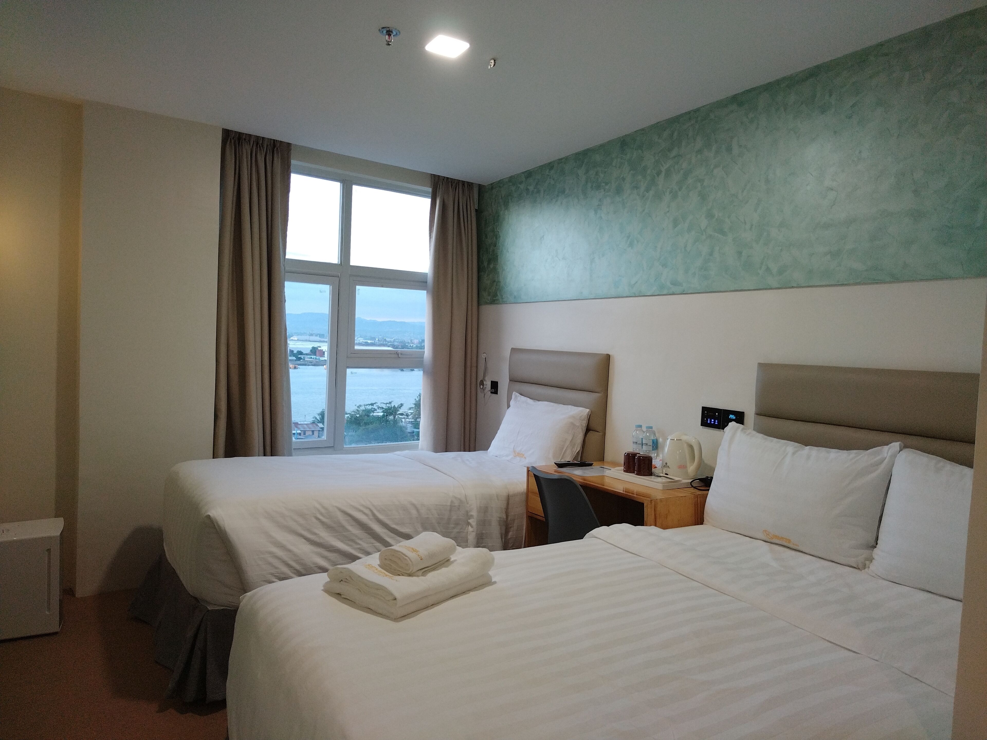 A hotel room with two beds, a desk, a chair, a nightstand with a phone and a kettle, and a view of the sea through a large window.