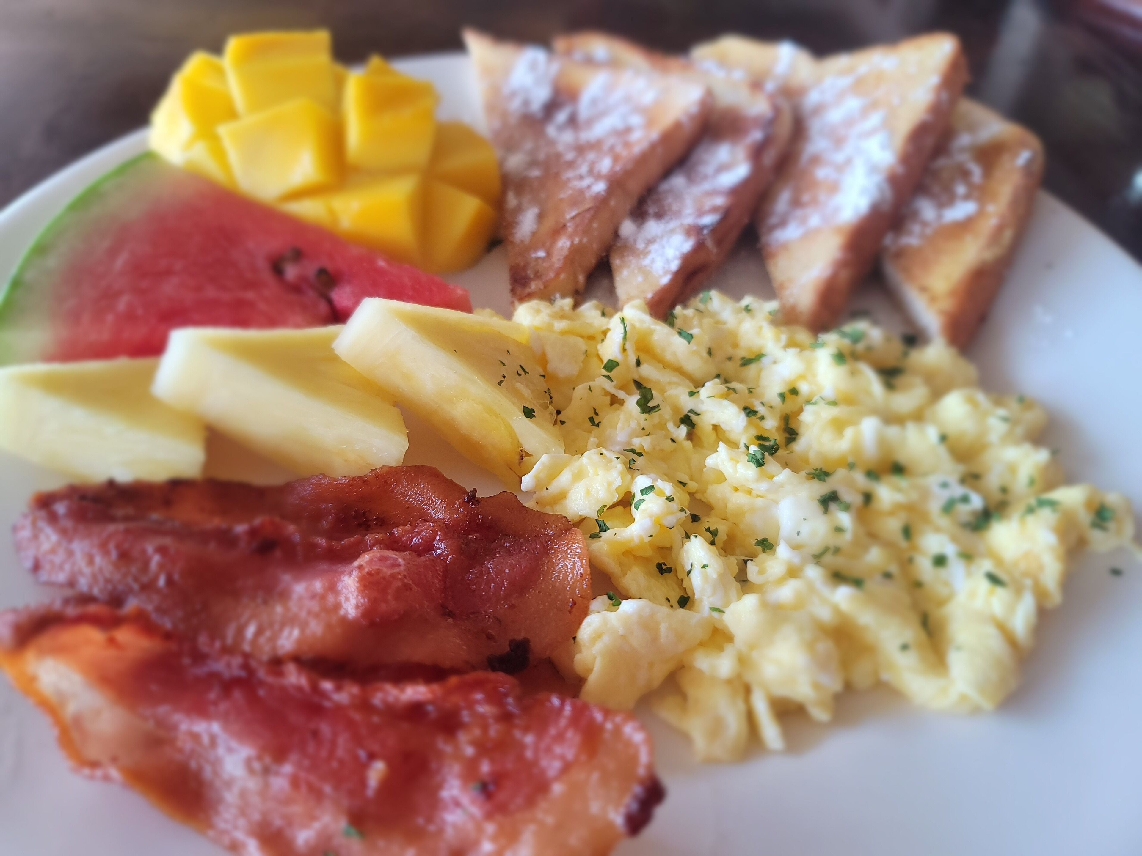 Un plato de desayuno con tostadas, huevos revueltos, tocino, piña y sandía.