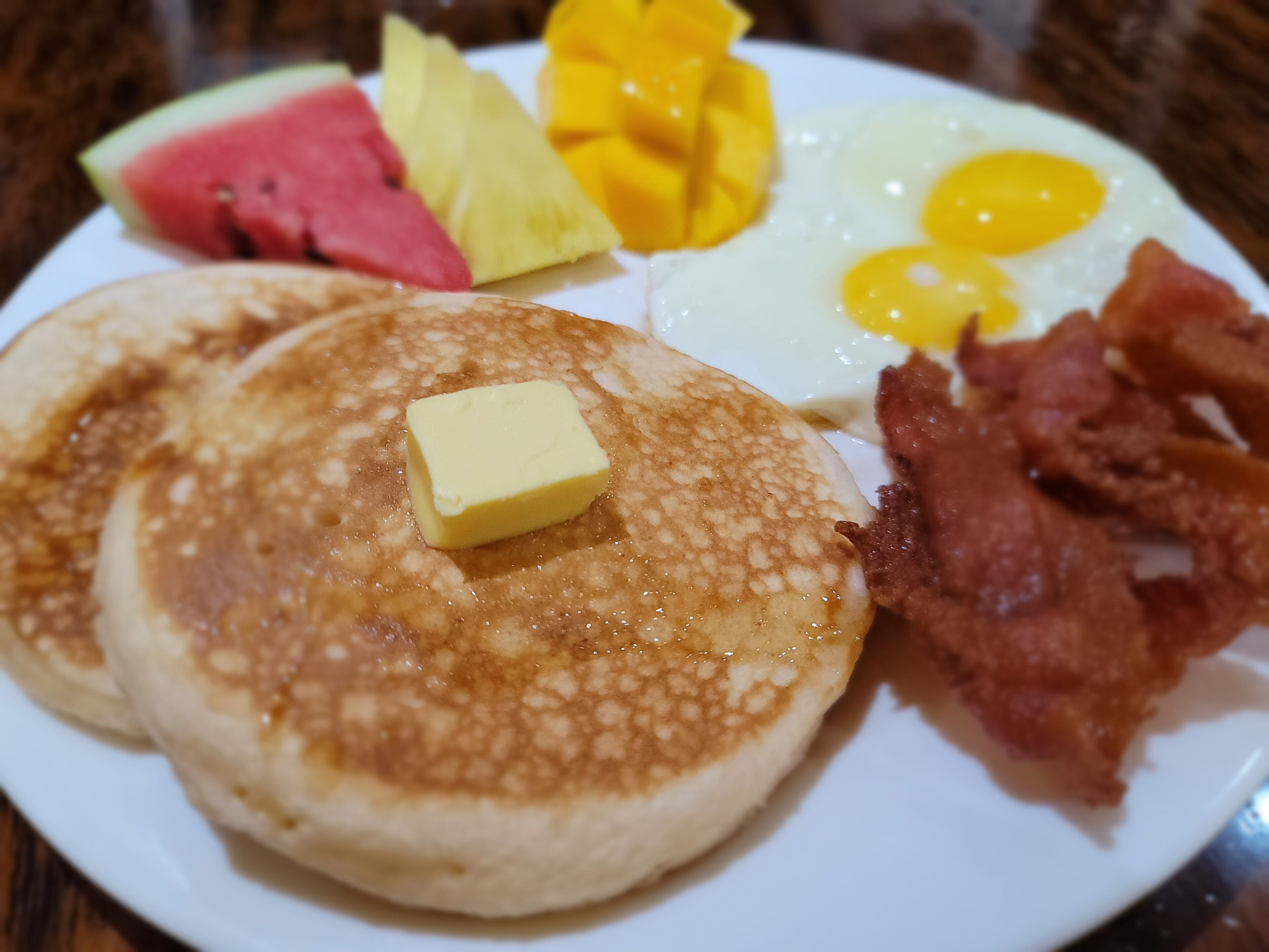 A breakfast plate with pancakes, eggs, bacon, butter, and fruit.