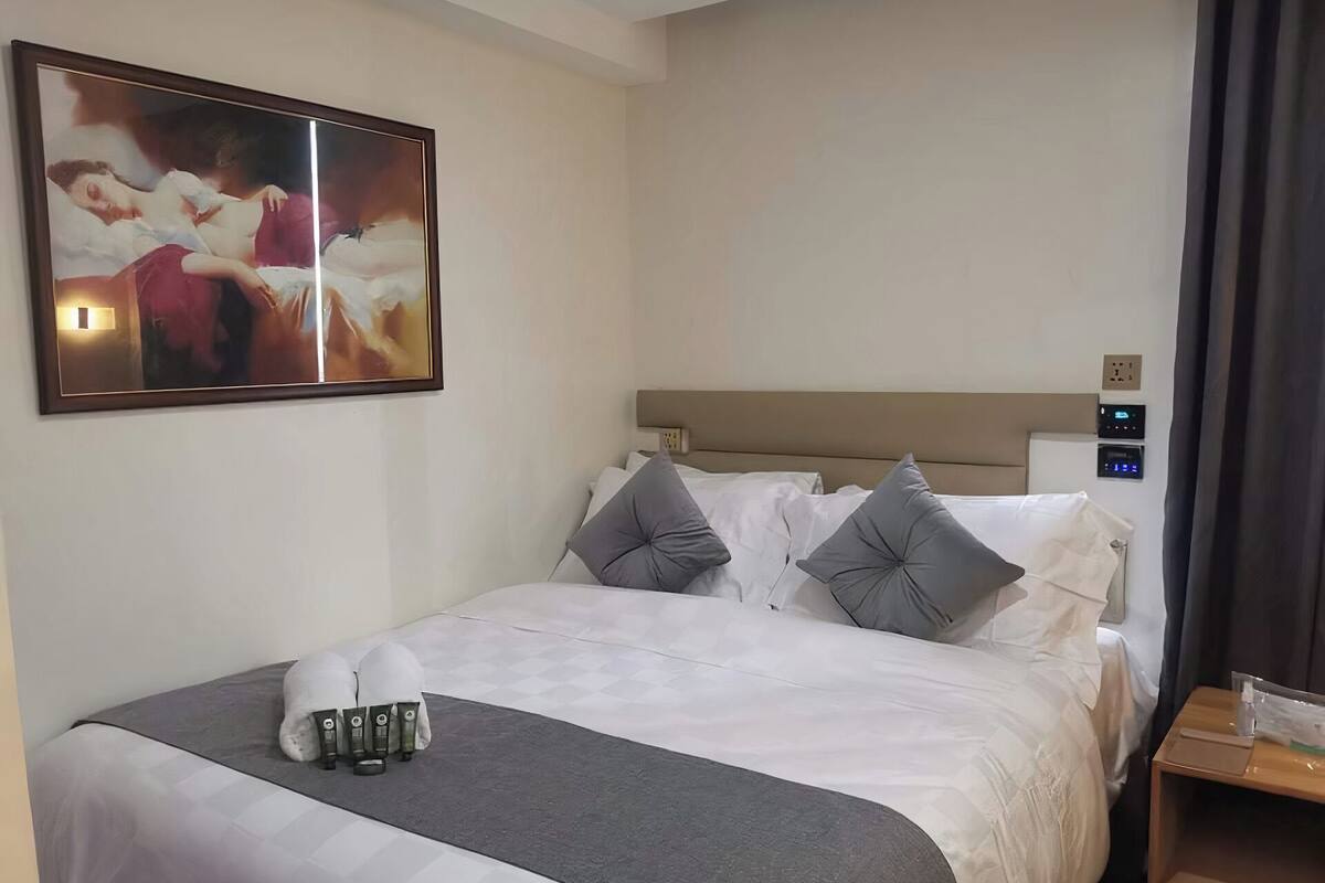 Une chambre d’hôtel comprenant un lit, des oreillers, une table de chevet, une commode, un panneau de commande fixé au mur et un tableau encadré au-dessus du lit.