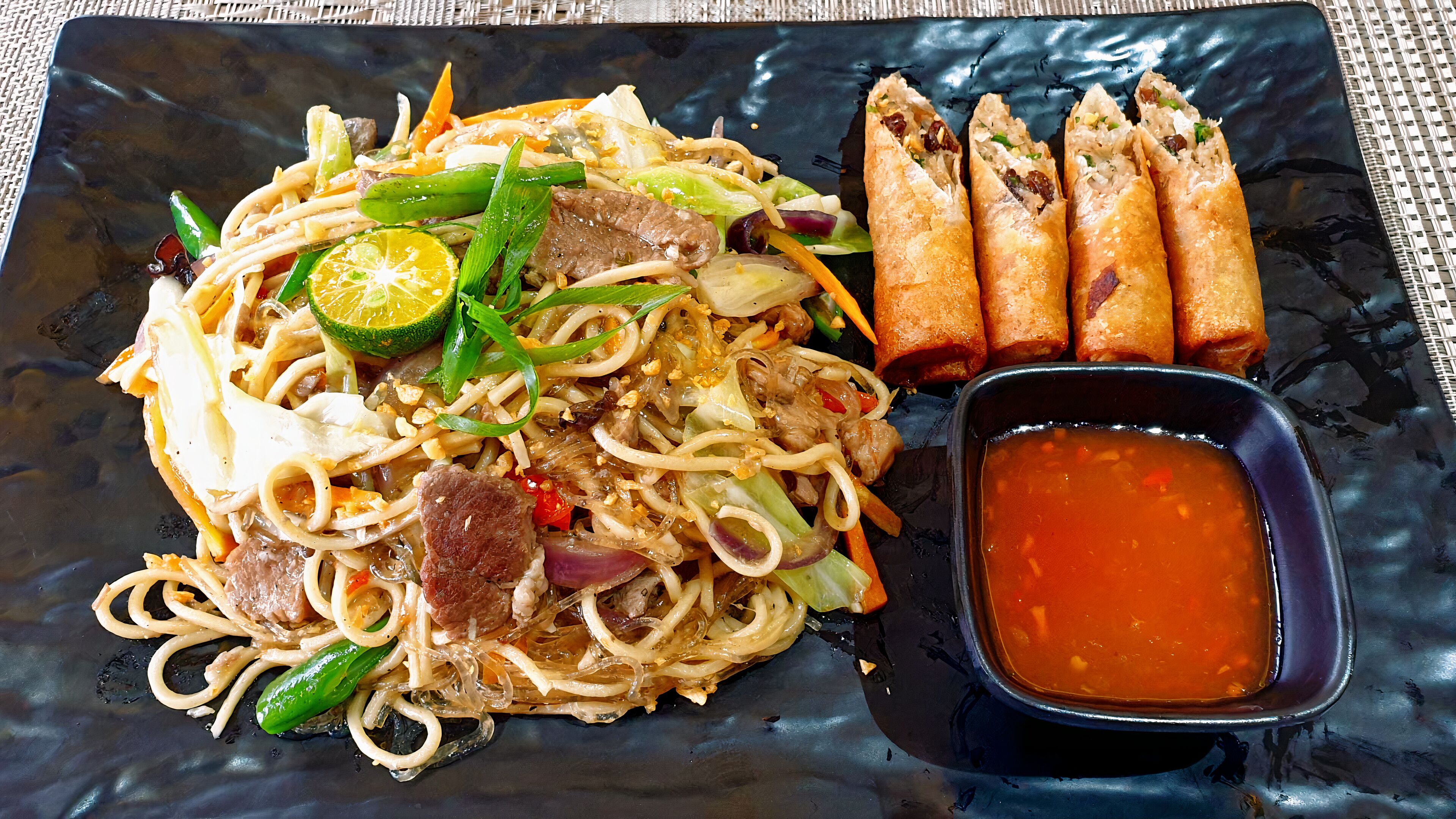 A plate of noodles with vegetables and meat, accompanied by spring rolls and a side of sauce.