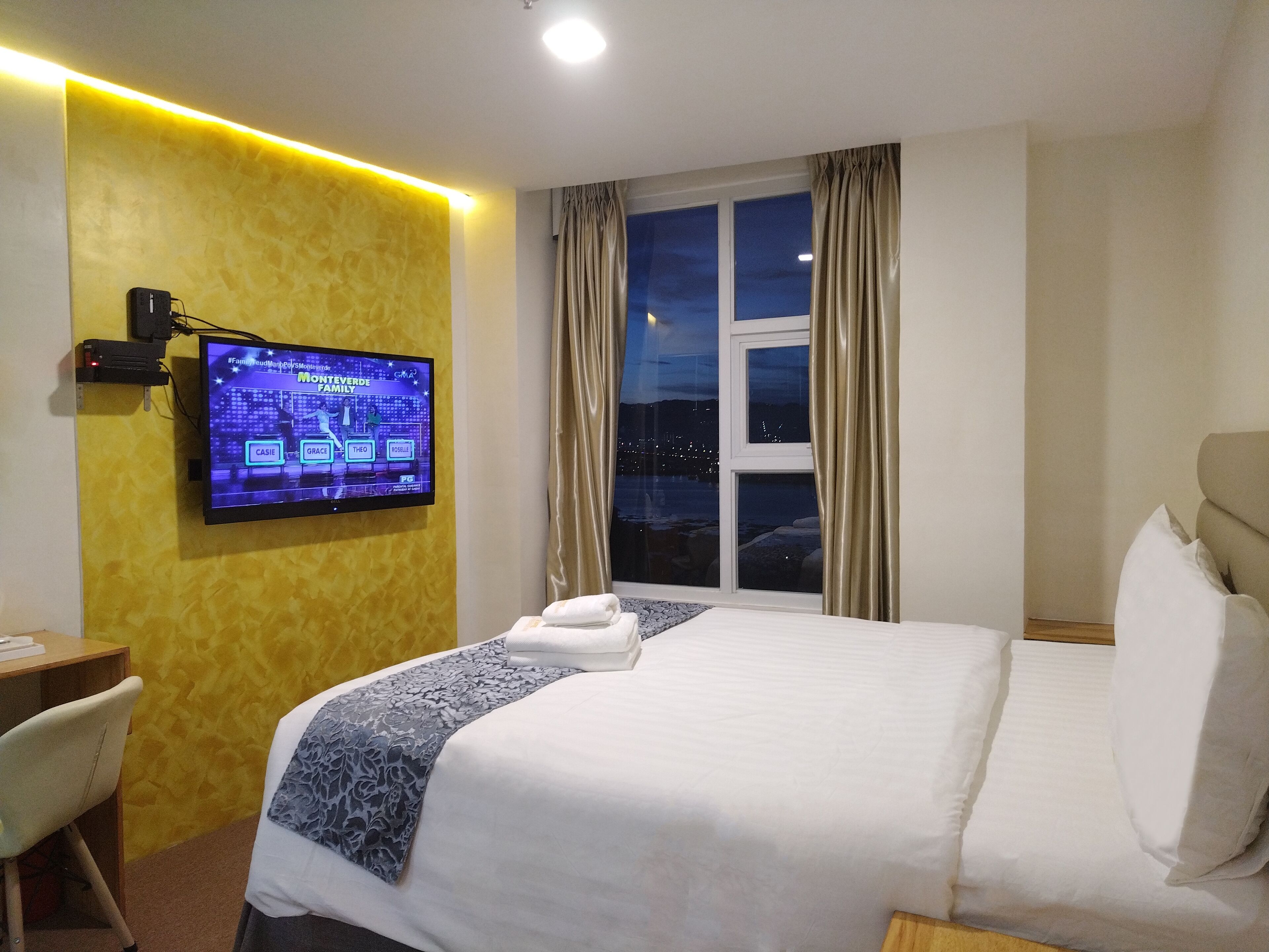 A hotel room with a bed, a desk, a television displaying a game show, and a window with curtains.