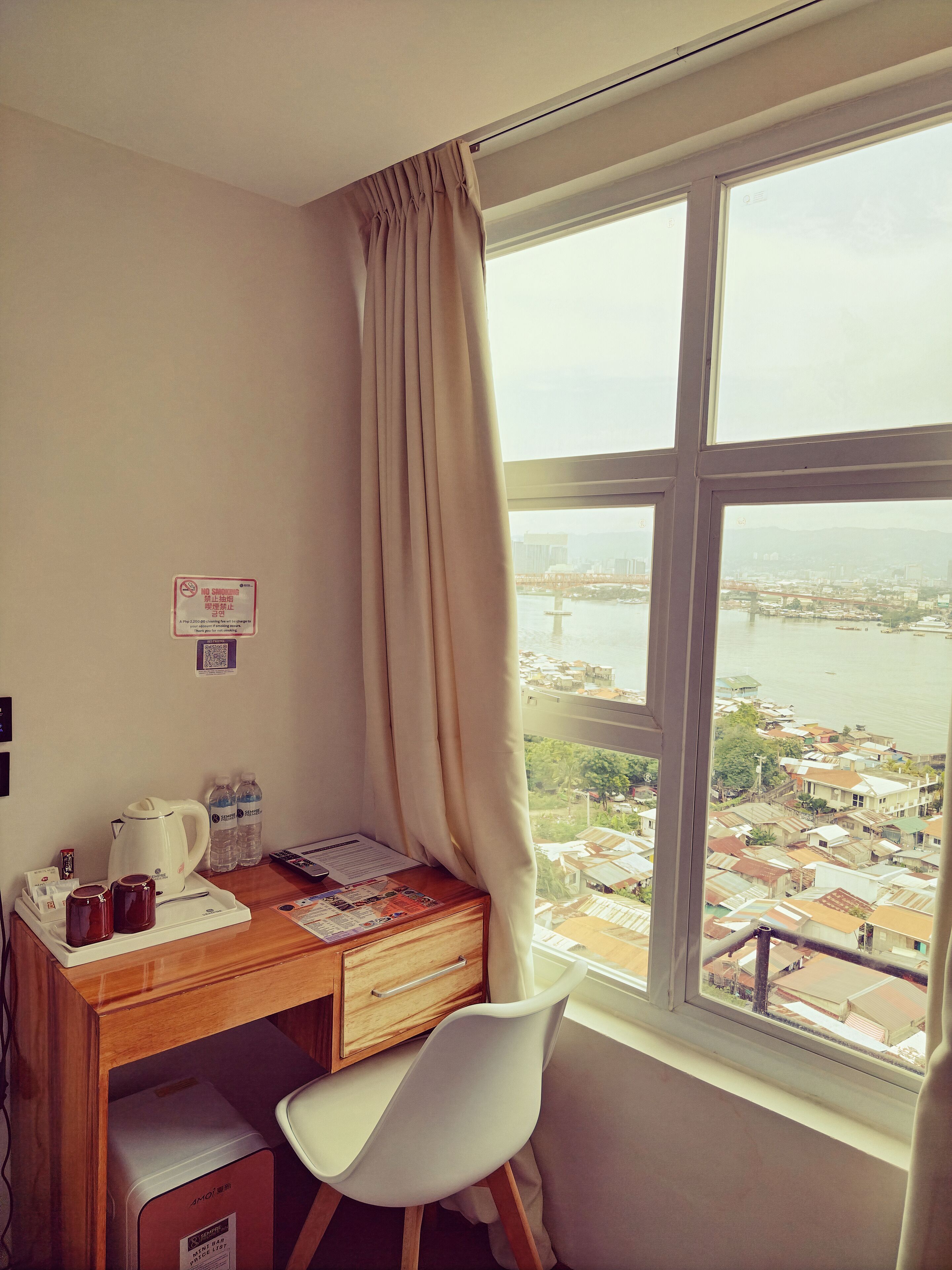 A hotel room with a desk, chair, and a view of a cityscape through a large window.