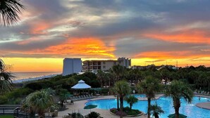 A resort pool area with a sunset view, palm trees, and a large building in the background. - Modern Coastal Retreat Near Miramar Beach With EV Charging Capablities (Destin)