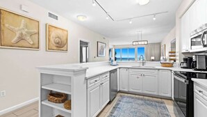 A modern kitchen with white cabinetry, a built-in dishwasher, and a large island with a sink. There are framed artworks on the wall and a view of the ocean through the windows. - Modern Coastal Retreat Near Miramar Beach With EV Charging Capablities (Destin)