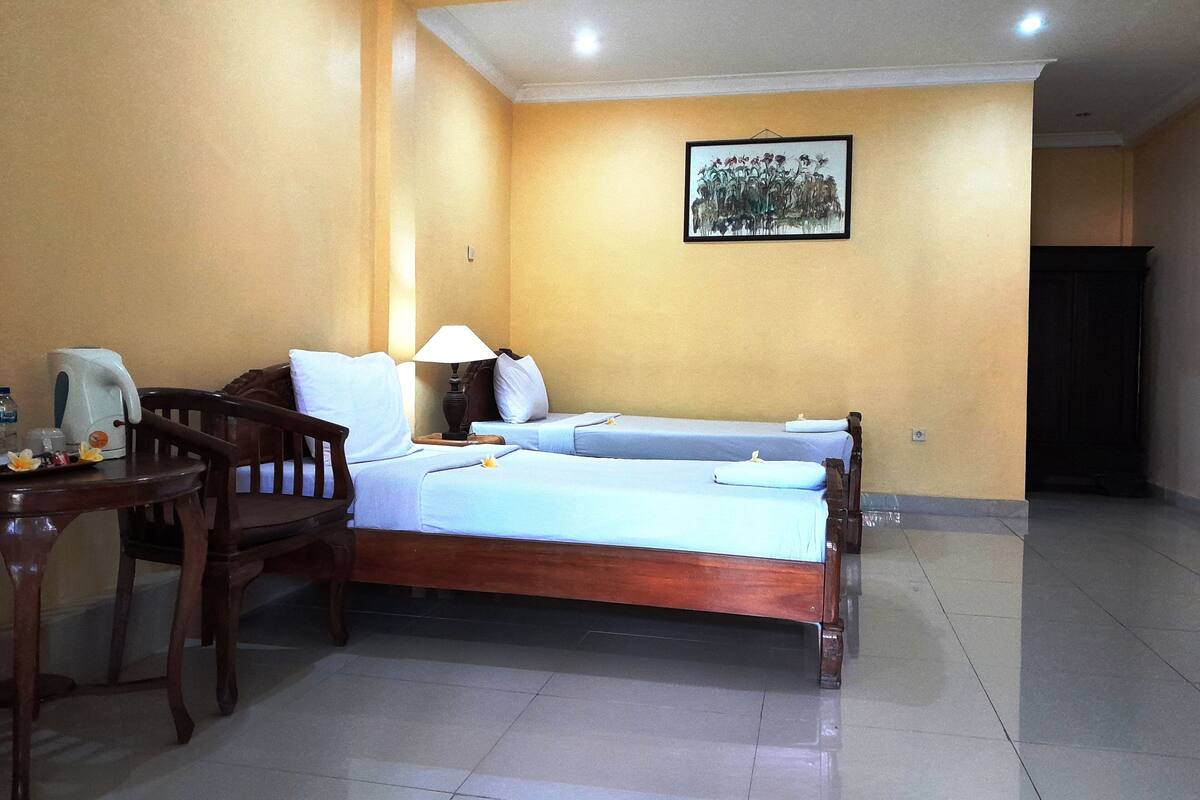 Una habitación de hotel con dos camas individuales, una silla de madera, una mesa con hervidor y una lámpara fijada a la pared.