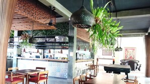 A restaurant interior with wooden tables and chairs, hanging plants, and a kitchen in the background. - Panorama Hotel Ubud (Ubud)