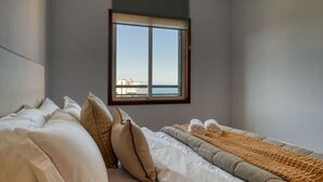 Habitación de hotel con cama, almohadas, manta y vista al mar a través de una ventana.