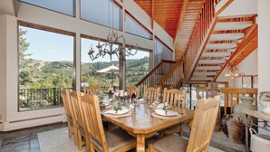 Un coin repas avec une table en bois dressée pour six personnes, entourée de chaises, un lustre au-dessus et une vue sur l’extérieur grâce à de grandes fenêtres.