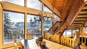 Une table à manger en bois, des chaises, un lustre et de grandes fenêtres donnant sur un paysage enneigé.