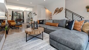 A modern living room with a grey sofa, wooden coffee table, and a staircase with a railing. - Luxury Townhome at Wasatch Springs - Black Rock Ridge + Ski Park City (Heber City)