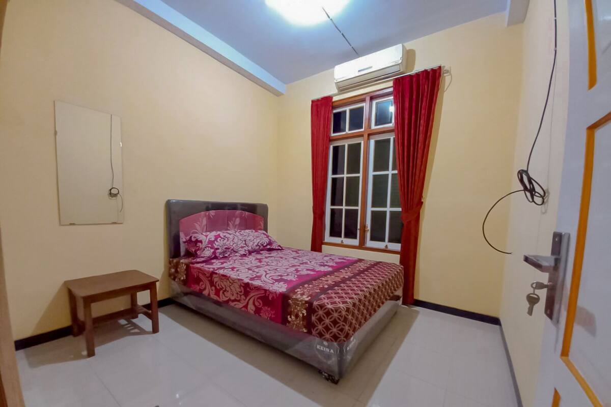 A bedroom with a bed, a small table, a window with curtains, an air conditioner, and a door.