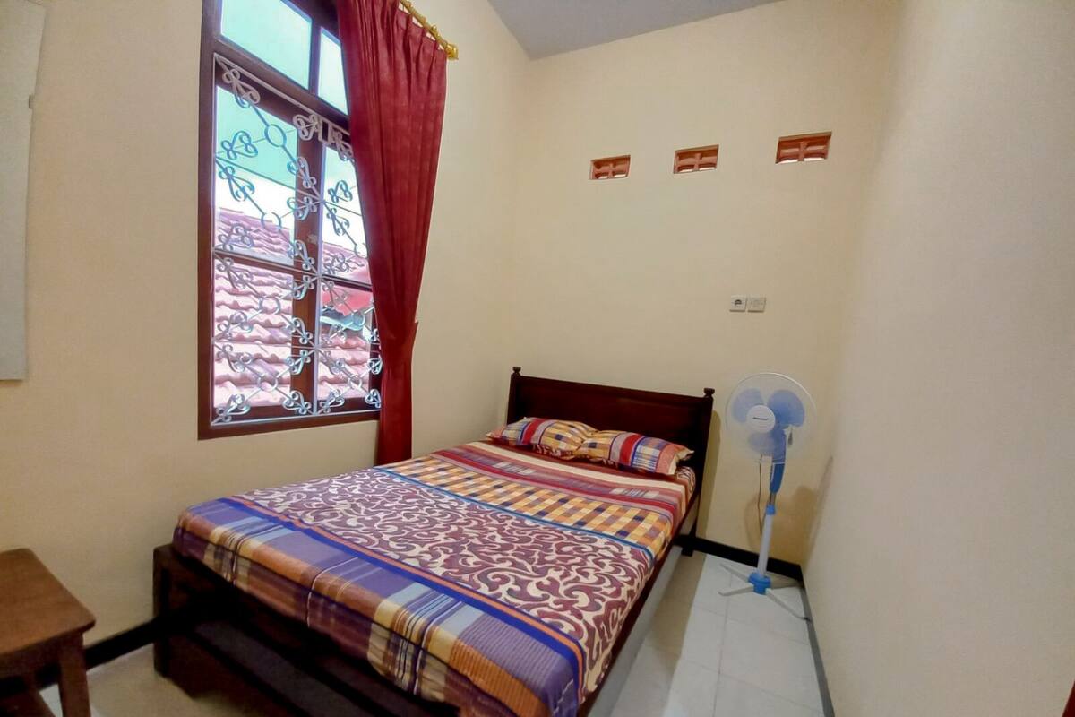 A bedroom with a bed, a window with curtains, a standing fan, and a small table.
