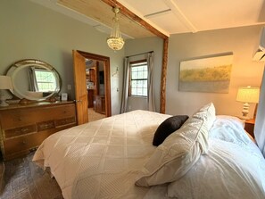 A bedroom with a bed, a dresser, a mirror, a chandelier, a window with curtains, and a painting on the wall. - Ligonier Creekside Cabin in Laurel Highlands (Ligonier)