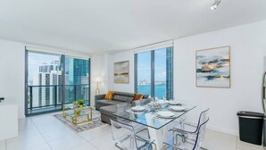 A modern living room with a glass dining table, grey sofa, and large windows offering city views.
