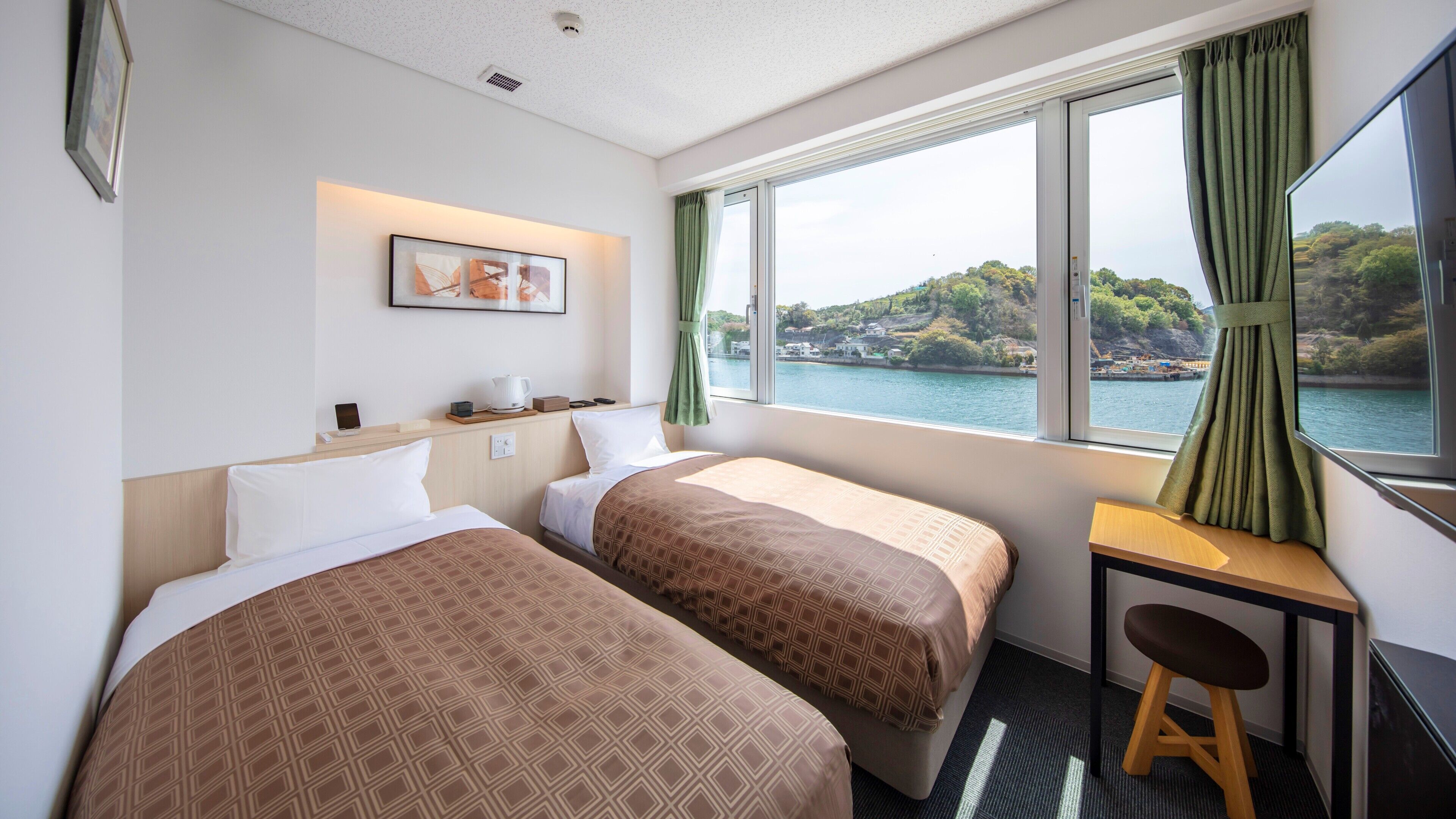 A hotel room with two beds, a desk, a chair, a window with a view of a body of water and hills, and a framed picture on the wall.