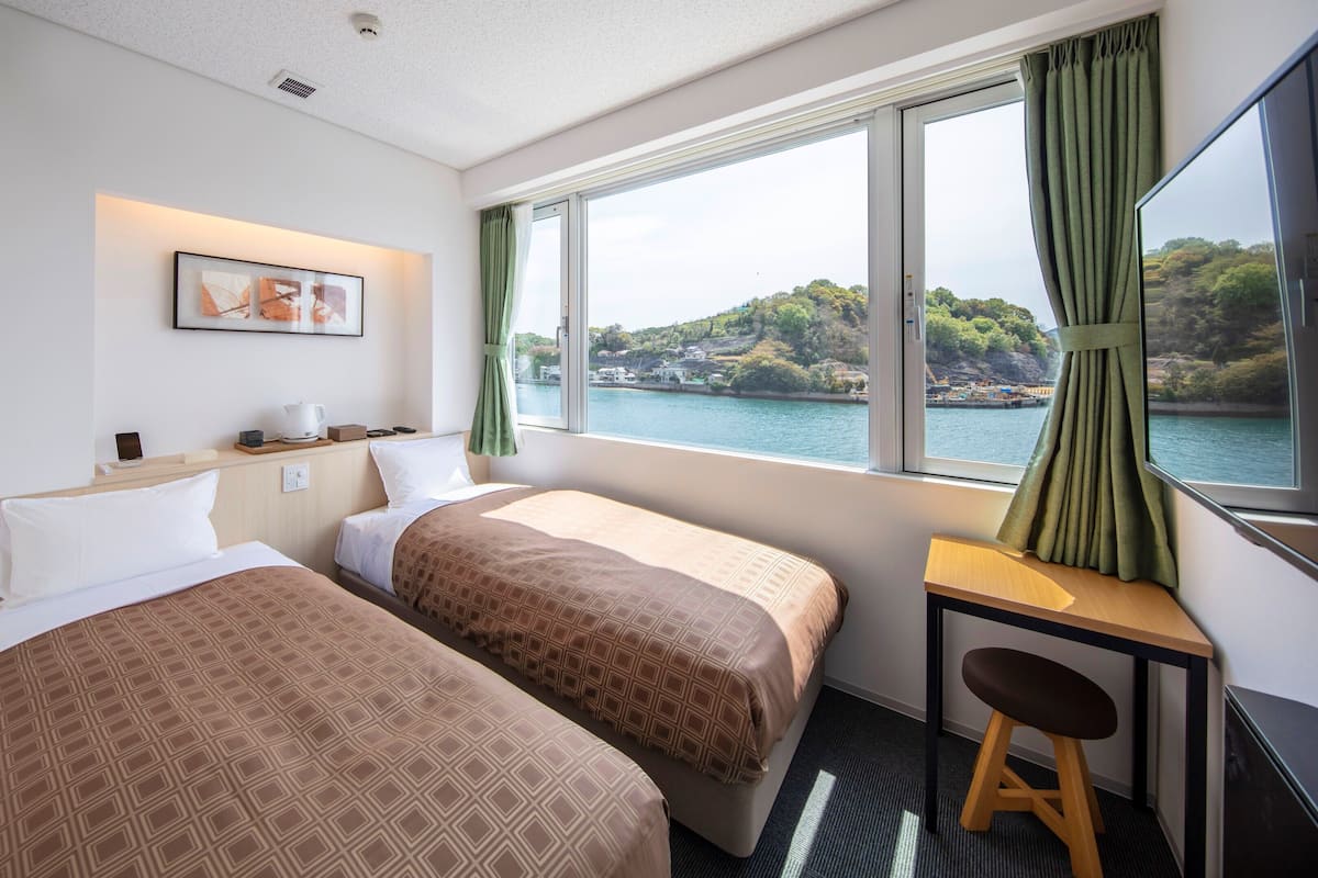 Une chambre d’hôtel avec deux lits, un bureau, une chaise, une fenêtre donnant sur un plan d’eau et des collines, et un tableau encadré au mur.