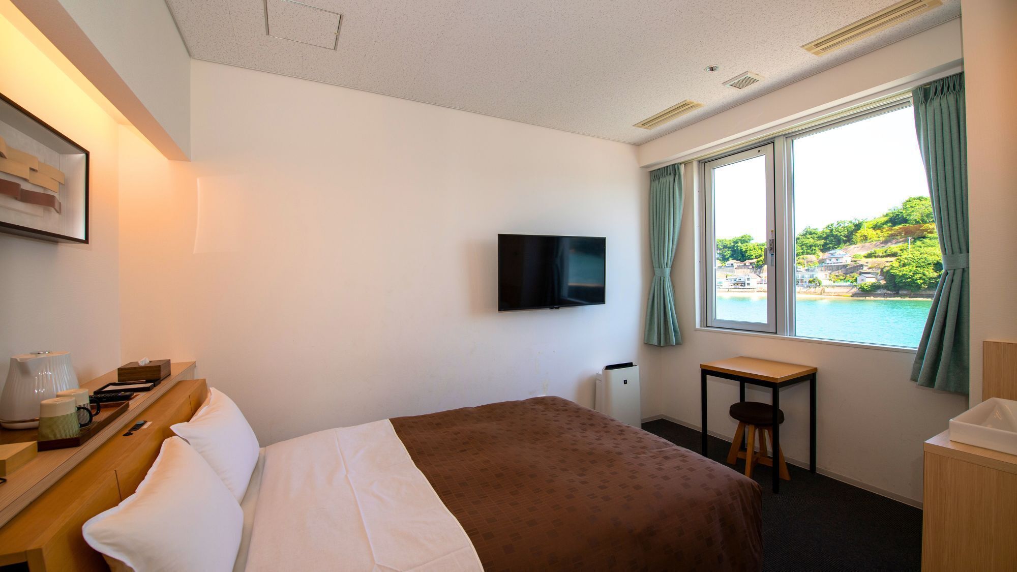 A hotel room with a bed, a desk, a chair, a TV, and a window with a view of a body of water and greenery.