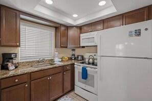 A kitchen with wooden cabinets, a white refrigerator, a microwave, a stove, and a window with blinds. - Bayfront Unit | Dock & Bay Views | Siesta Key (Sarasota)