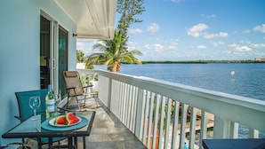 A balcony with a table set for two, overlooking a body of water with a palm tree and distant buildings.
