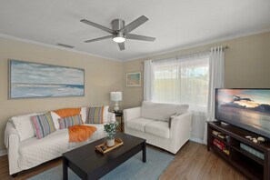 A living room with a ceiling fan, two white sofas, a coffee table, a TV stand, and a window with curtains.